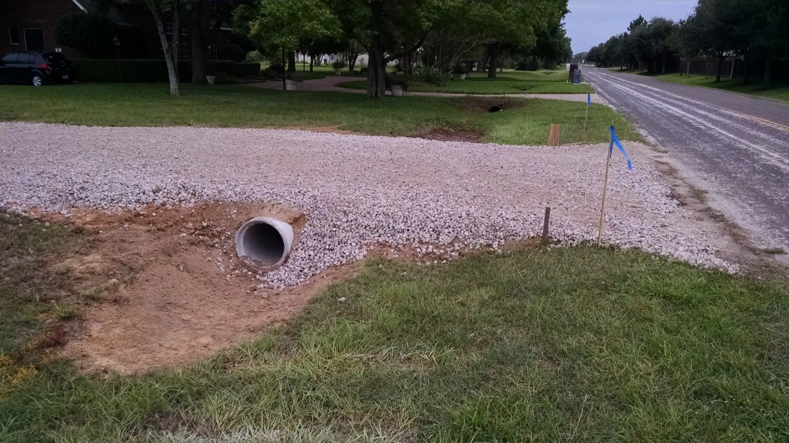 Plastic culvert pipe under a new gravel driveway apron beside a paved road, survey flags in the ditch